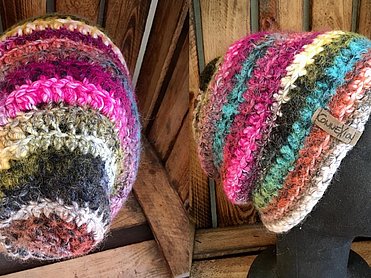 Handgefertigte Beanie-Mütze im Regenbogenmix mit bunten Querstreifen in Pink, Türkis, Erdtönen und Grau sowie Logo-Label.